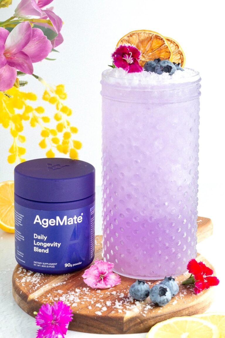 A glass of purple drink garnished with blueberries, edible flowers, and dried citrus slices sits next to a purple AgeMate Daily Longevity Blend container on a wooden board with flowers, lemon slices, and blueberries around.