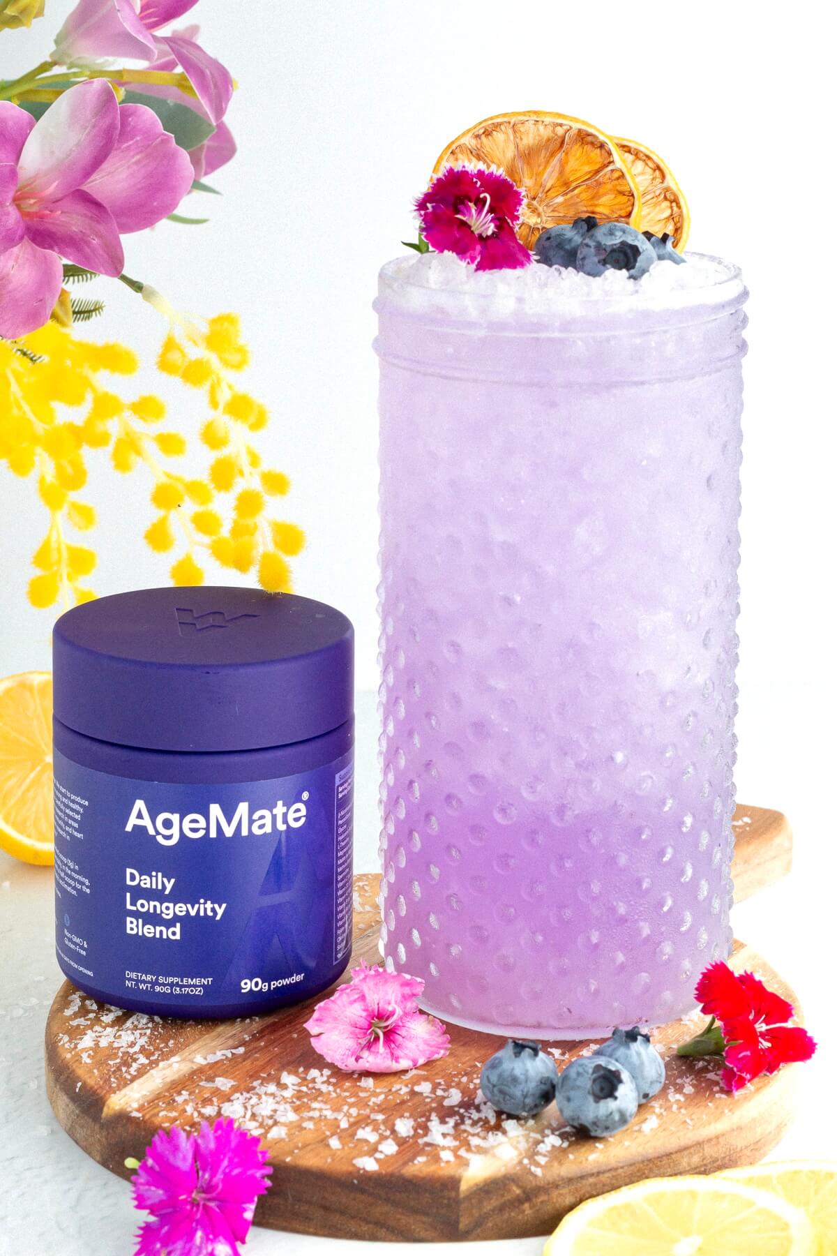 A glass of purple drink garnished with blueberries, edible flowers, and dried citrus slices sits next to a purple AgeMate Daily Longevity Blend container on a wooden board with flowers, lemon slices, and blueberries around.