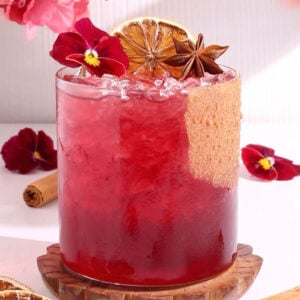 A vibrant red mocktail in a glass with crushed ice, garnished with a dried citrus slice, star anise, purple edible flowers, and a cinnamon stick, with more flowers and spices around the drink.