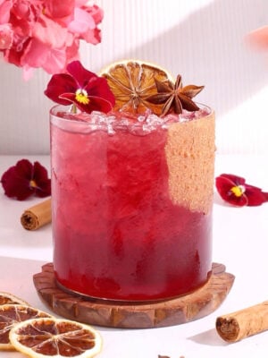 A vibrant red mocktail in a glass with crushed ice, garnished with a dried citrus slice, star anise, purple edible flowers, and a cinnamon stick, with more flowers and spices around the drink.