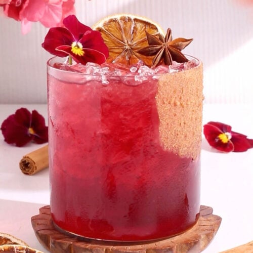 A vibrant red mocktail in a glass with crushed ice, garnished with a dried citrus slice, star anise, purple edible flowers, and a cinnamon stick, with more flowers and spices around the drink.