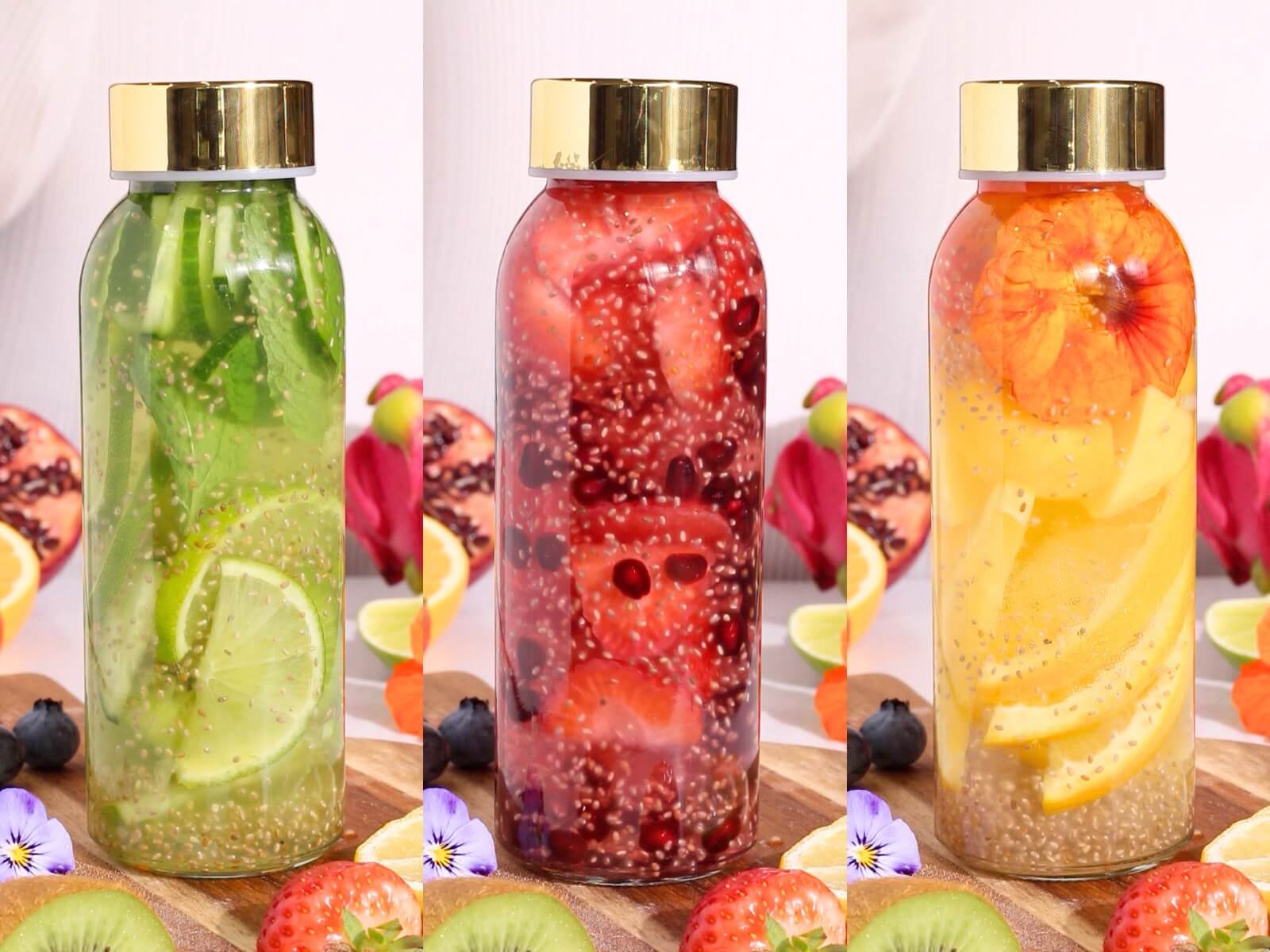 Three glass bottles with gold caps, each filled with fruit-infused water. The bottles contain slices of green lime and kiwi, red strawberries and pomegranate, and orange citrus with yellow fruit, all with chia seeds. Fruits are scattered around.