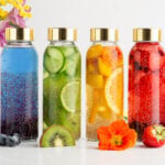 Six glass bottles with gold lids contain colorful infused waters with fruits and chia seeds. From left to right: blueberry, blue, cucumber, citrus, red berry, and pink mixtures. Flowers and fruit slices are in front and behind.