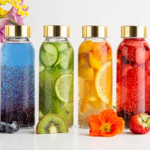 Six glass bottles with gold lids contain colorful infused waters with fruits and chia seeds. From left to right: blueberry, blue, cucumber, citrus, red berry, and pink mixtures. Flowers and fruit slices are in front and behind.