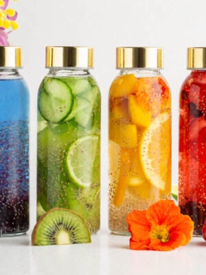 Six glass bottles with gold lids contain colorful infused waters with fruits and chia seeds. From left to right: blueberry, blue, cucumber, citrus, red berry, and pink mixtures. Flowers and fruit slices are in front and behind.