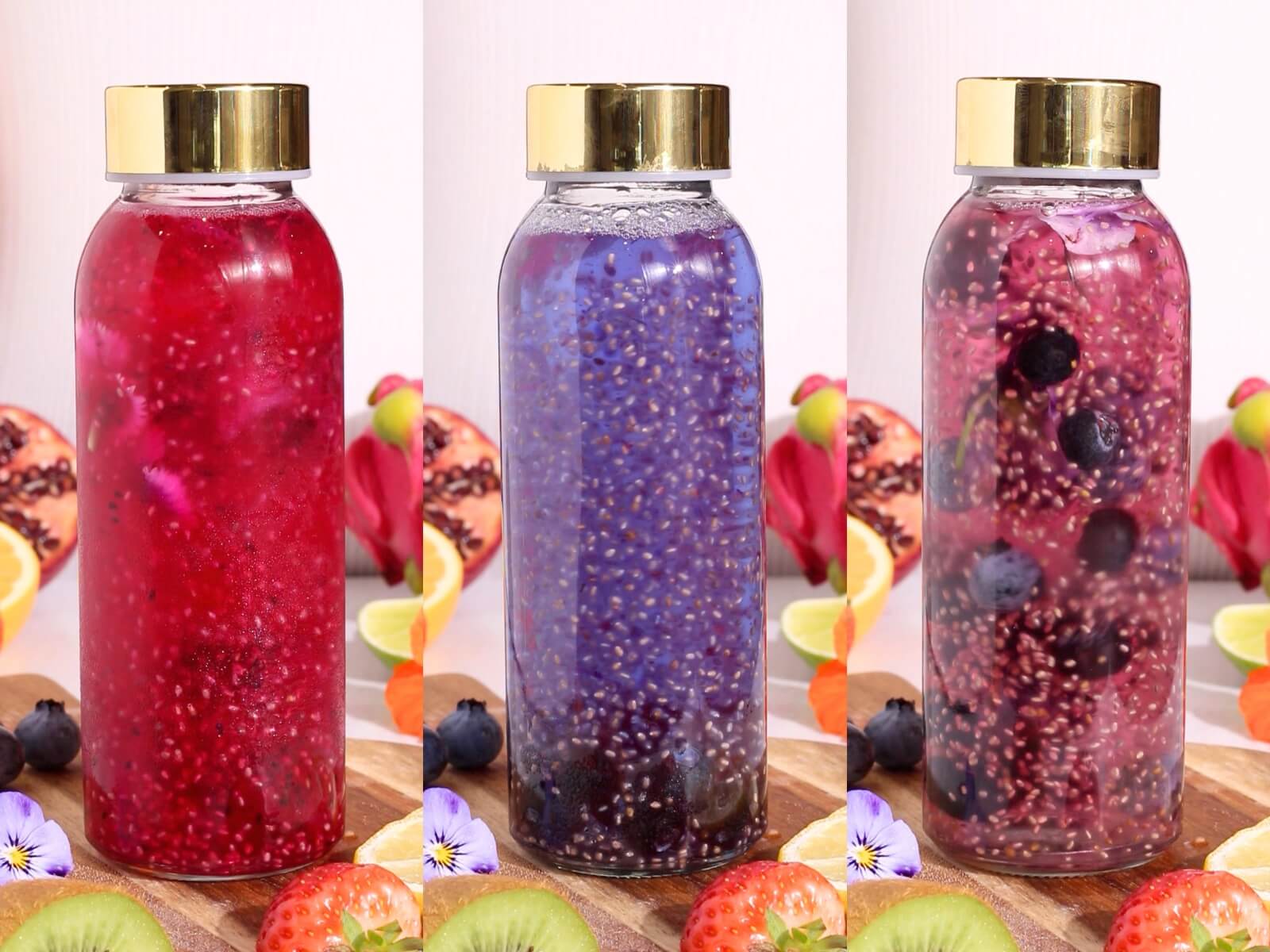 Three glass bottles with gold caps are filled with colorful chia seed drinks—red, purple, and pink—surrounded by sliced kiwi, strawberries, lemon, pomegranate, blueberries, and edible flowers on a wooden surface.