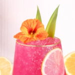A vibrant pink drink with ice is garnished with a slice of lemon, a bright orange edible flower, and two green leaves. The background is light and minimal, with lemon slices visible around the glass.