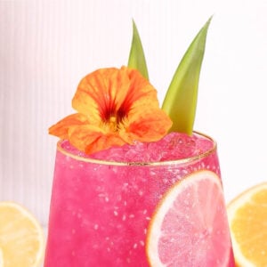 A vibrant pink drink with ice is garnished with a slice of lemon, a bright orange edible flower, and two green leaves. The background is light and minimal, with lemon slices visible around the glass.