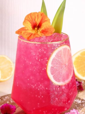 A hot pink punch garnished with a lemon slice, orange flower, and green leaves, served in a faceted glass with ice. Lemon halves and pink flowers are in the background.