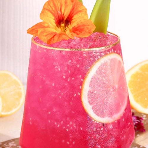 A hot pink punch garnished with a lemon slice, orange flower, and green leaves, served in a faceted glass with ice. Lemon halves and pink flowers are in the background.