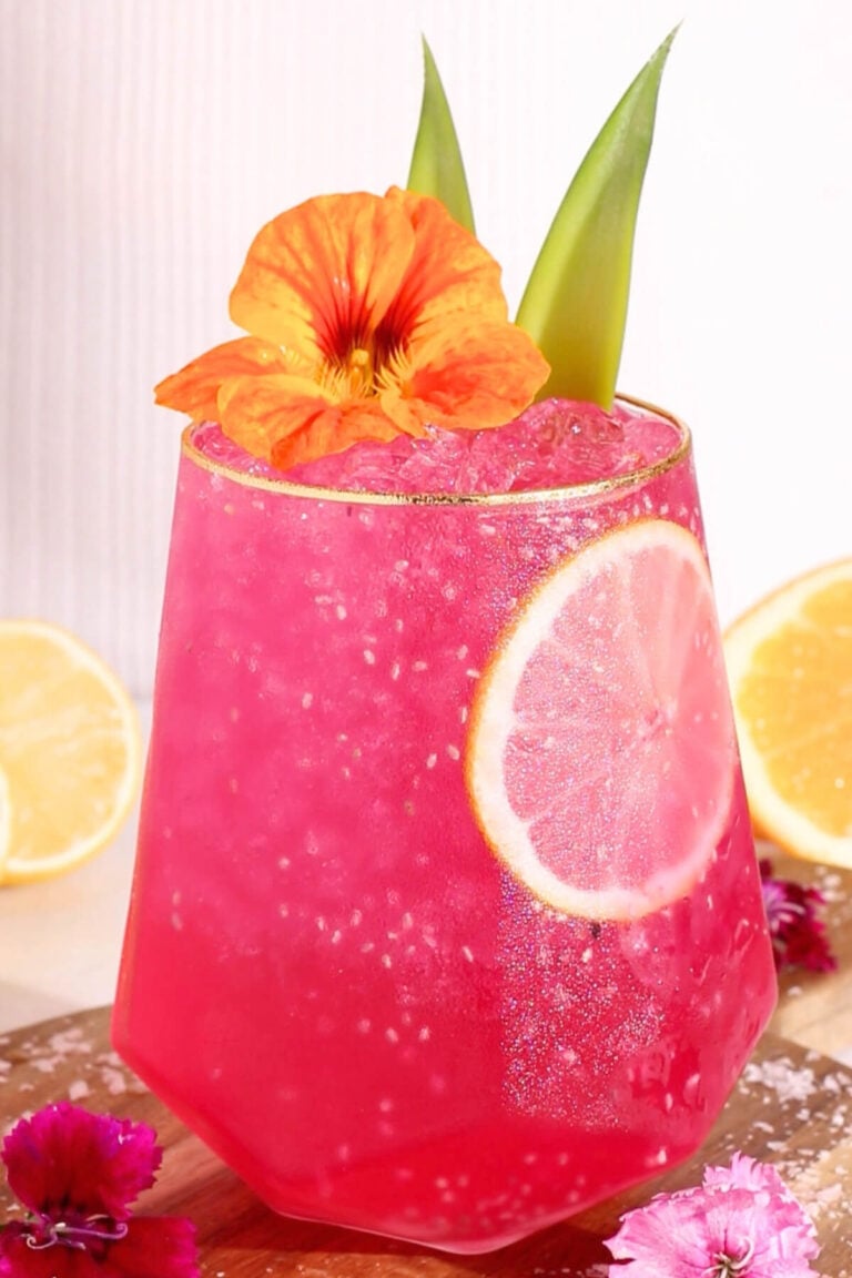 A hot pink punch garnished with a lemon slice, orange flower, and green leaves, served in a faceted glass with ice. Lemon halves and pink flowers are in the background.