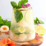 A glass of refreshing drink with ice, lychee fruit, lime slices, mint leaves, and pink edible flowers, set on a wooden board with more mint, lime slices, and flowers around.