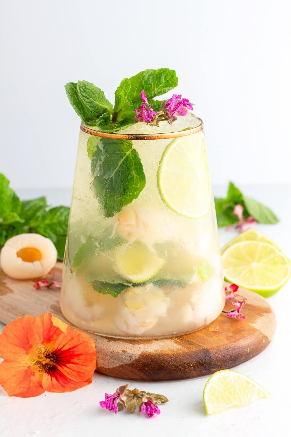 A glass of refreshing drink with ice, lychee fruit, lime slices, mint leaves, and pink edible flowers, set on a wooden board with more mint, lime slices, and flowers around.