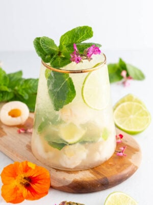 A refreshing drink with lime slices, lychee pieces, mint leaves, and ice in a clear glass, garnished with pink flowers and more mint. A sliced lime and colorful flowers are on a wooden board nearby.