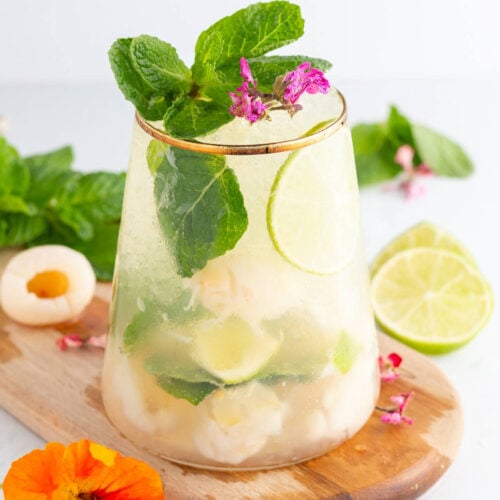 A refreshing drink with lime slices, lychee pieces, mint leaves, and ice in a clear glass, garnished with pink flowers and more mint. A sliced lime and colorful flowers are on a wooden board nearby.
