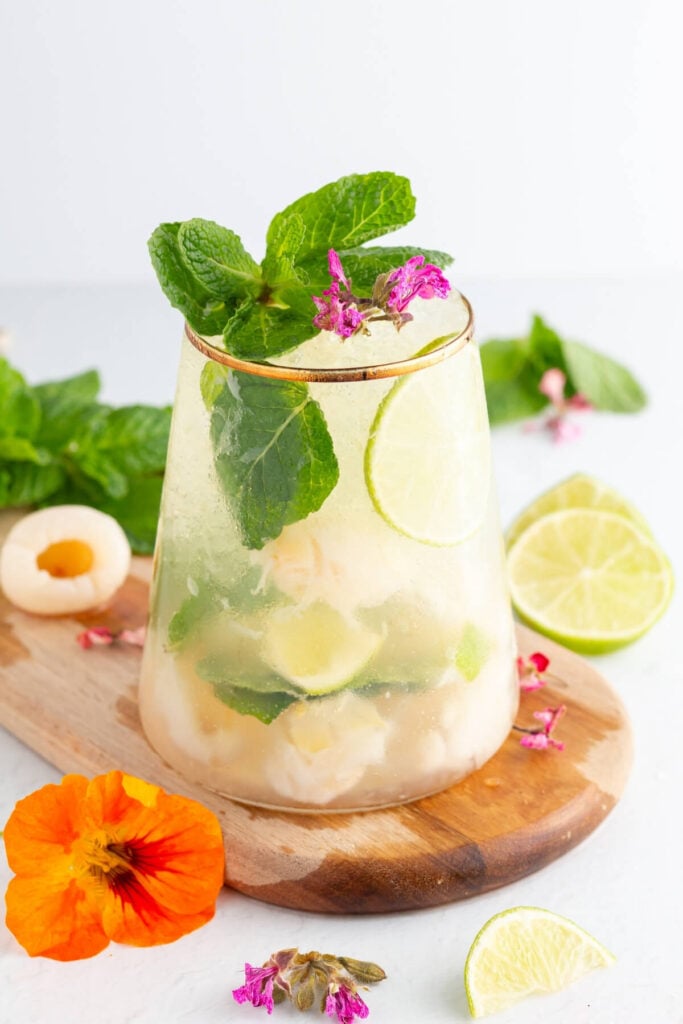 A refreshing drink with lime slices, lychee pieces, mint leaves, and ice in a clear glass, garnished with pink flowers and more mint. A sliced lime and colorful flowers are on a wooden board nearby.