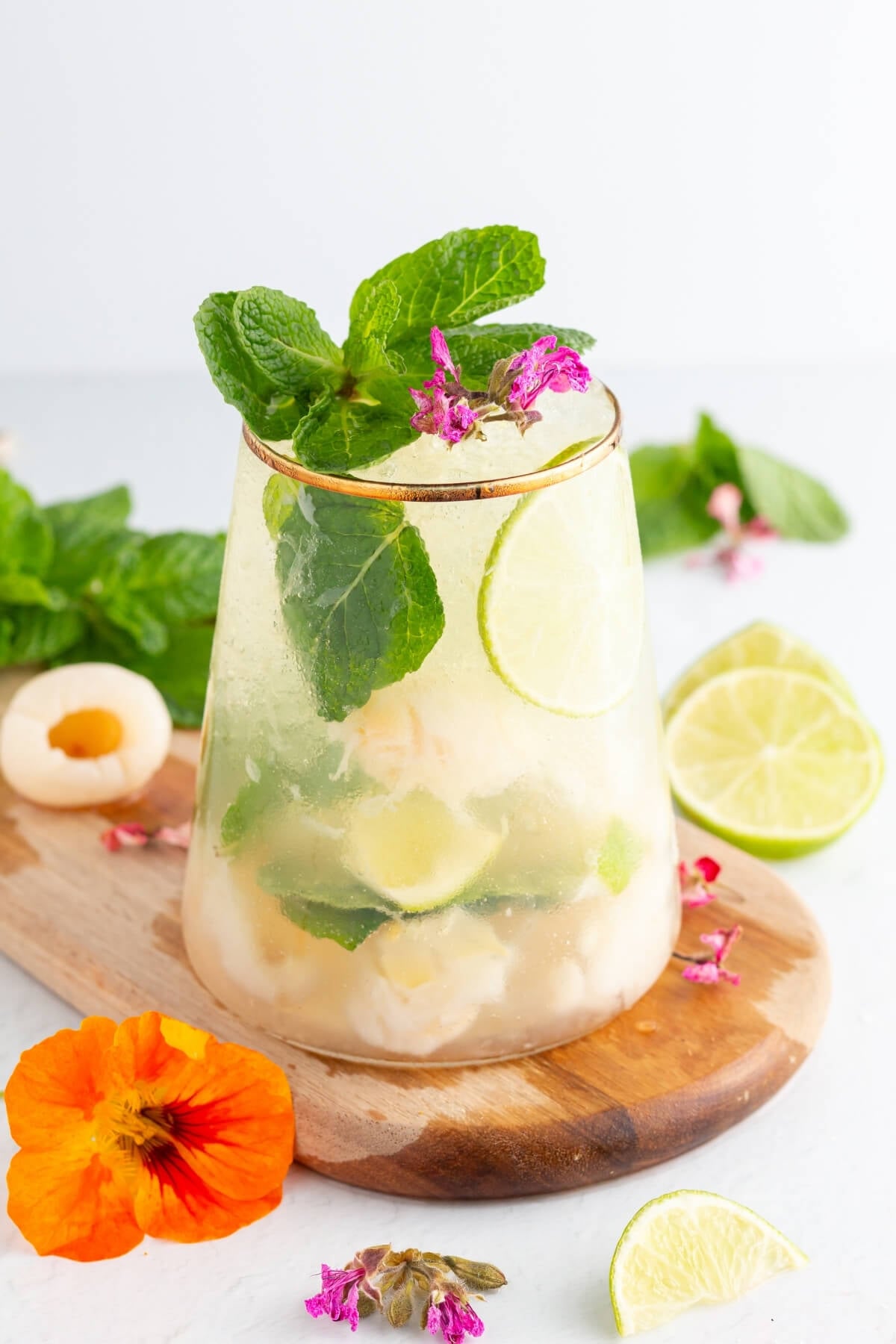 A refreshing drink with lime slices, lychee pieces, mint leaves, and ice in a clear glass, garnished with pink flowers and more mint. A sliced lime and colorful flowers are on a wooden board nearby.