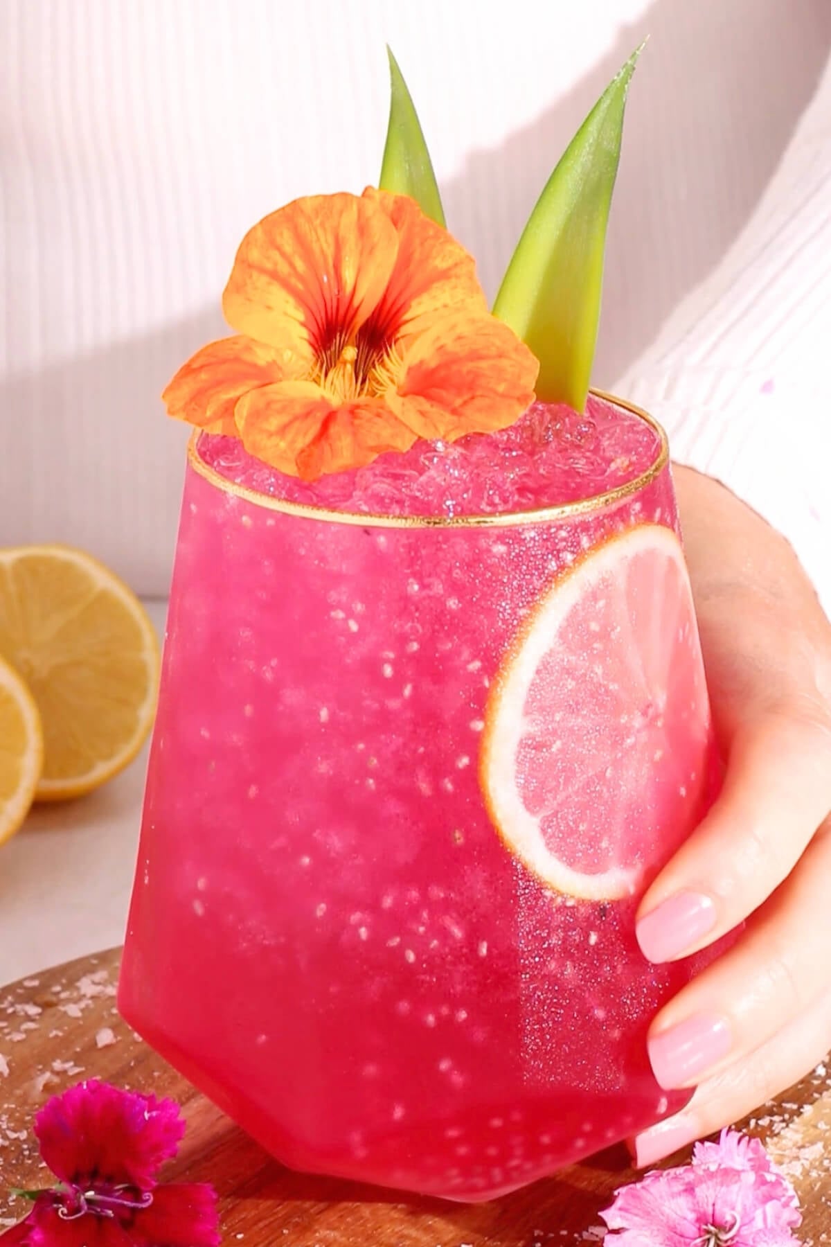 A vibrant pink drink with crushed ice in a glass, garnished with a lemon slice, green leaves, and an orange edible flower. A hand with pink nails holds the glass; lemons and pink flowers are visible in the background.
