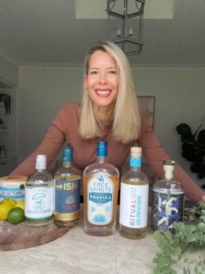 A smiling woman with long blonde hair stands behind a table displaying five bottles of non-alcoholic tequila alternatives, a container of sea salt flakes, and some limes in a bright, modern kitchen.