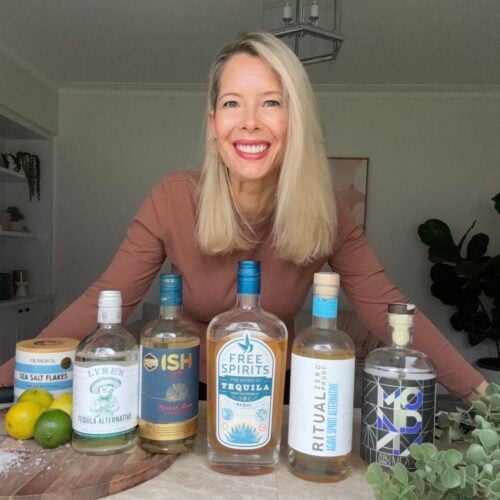 A smiling woman with long blonde hair stands behind a table displaying five bottles of non-alcoholic tequila alternatives, a container of sea salt flakes, and some limes in a bright, modern kitchen.