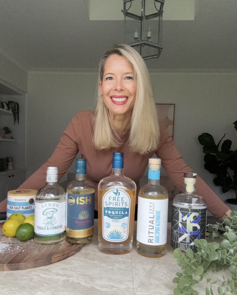 A smiling woman with long blonde hair stands behind a table displaying five bottles of non-alcoholic tequila alternatives, a container of sea salt flakes, and some limes in a bright, modern kitchen.