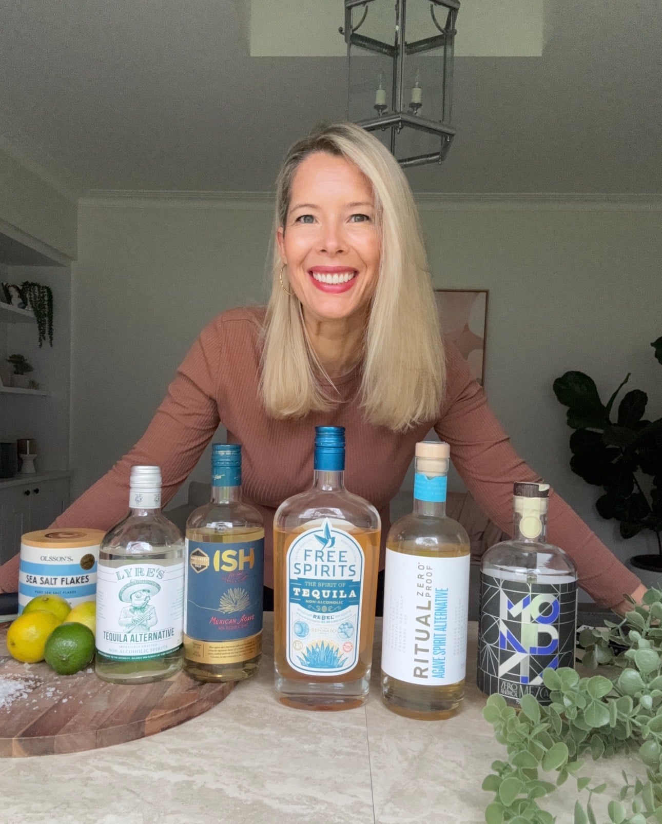 A smiling woman with long blonde hair stands behind a table displaying five bottles of non-alcoholic tequila alternatives, a container of sea salt flakes, and some limes in a bright, modern kitchen.
