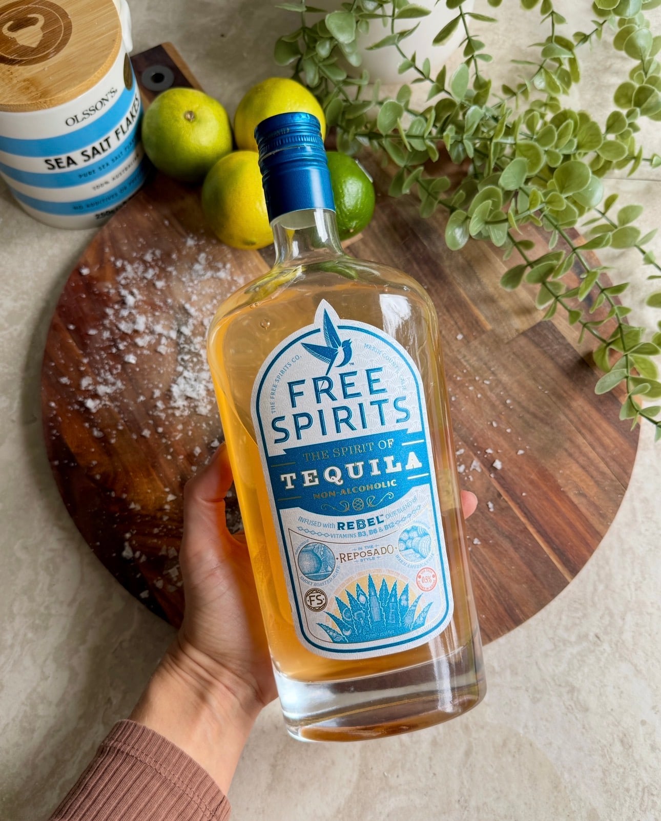 A hand holds a bottle of Free Spirits non-alcoholic tequila over a wooden board with sea salt, limes, and a plant in the background.