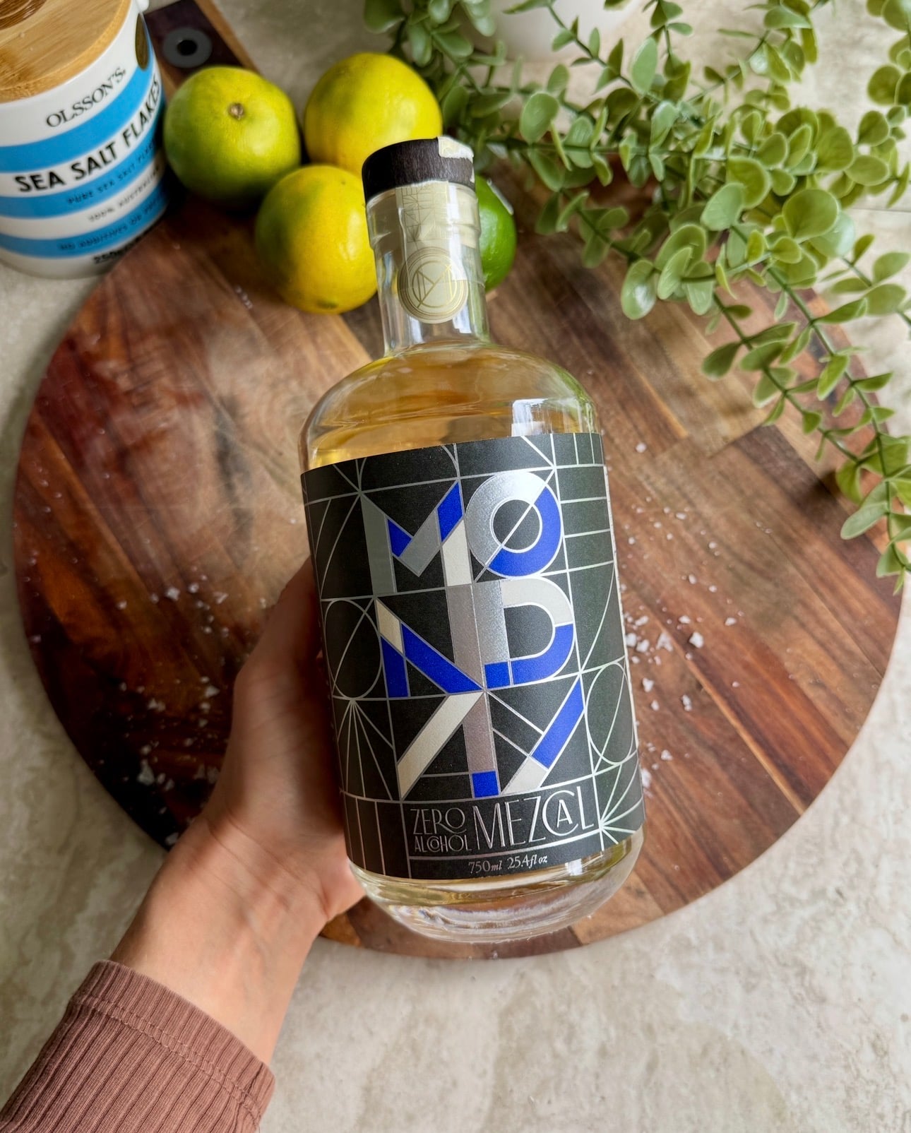 A hand holds a bottle labeled Monday Mezcal over a round wooden board with limes, sea salt, and green leaves nearby.