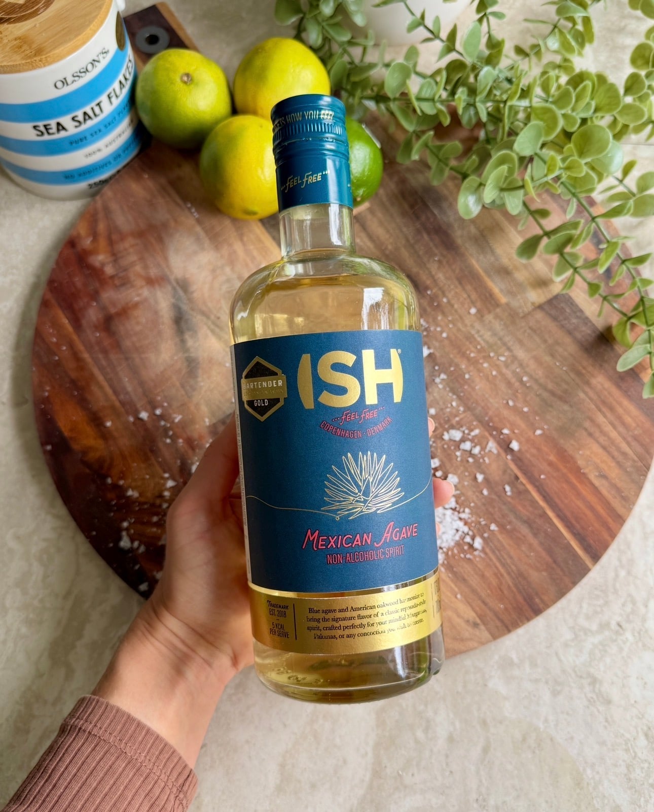 A hand holds a bottle of ISH Mexican Agave non-alcoholic spirit on a wooden board, with limes, a plant, and sea salt flakes in the background.