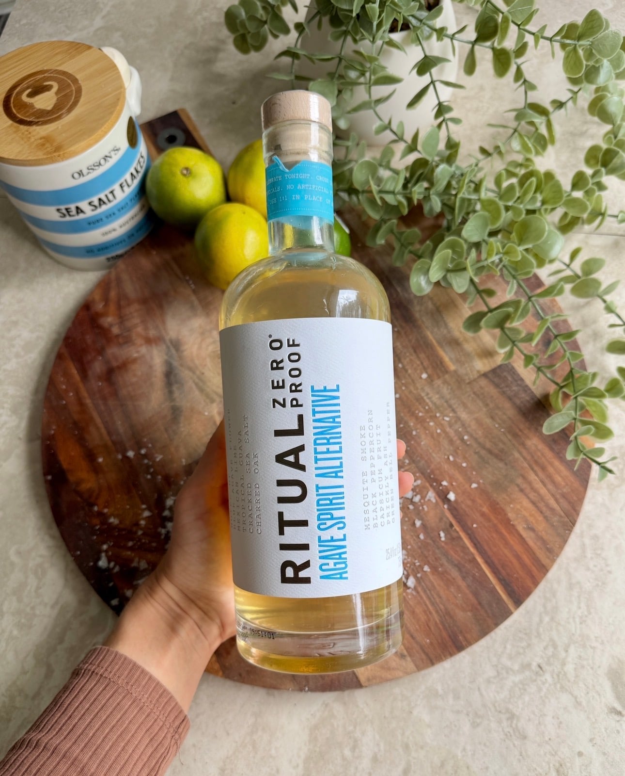 A hand holds a bottle of Ritual Zero Proof Agave Spirit Alternative above a wooden board with limes, a container of sea salt flakes, and green plant leaves nearby.