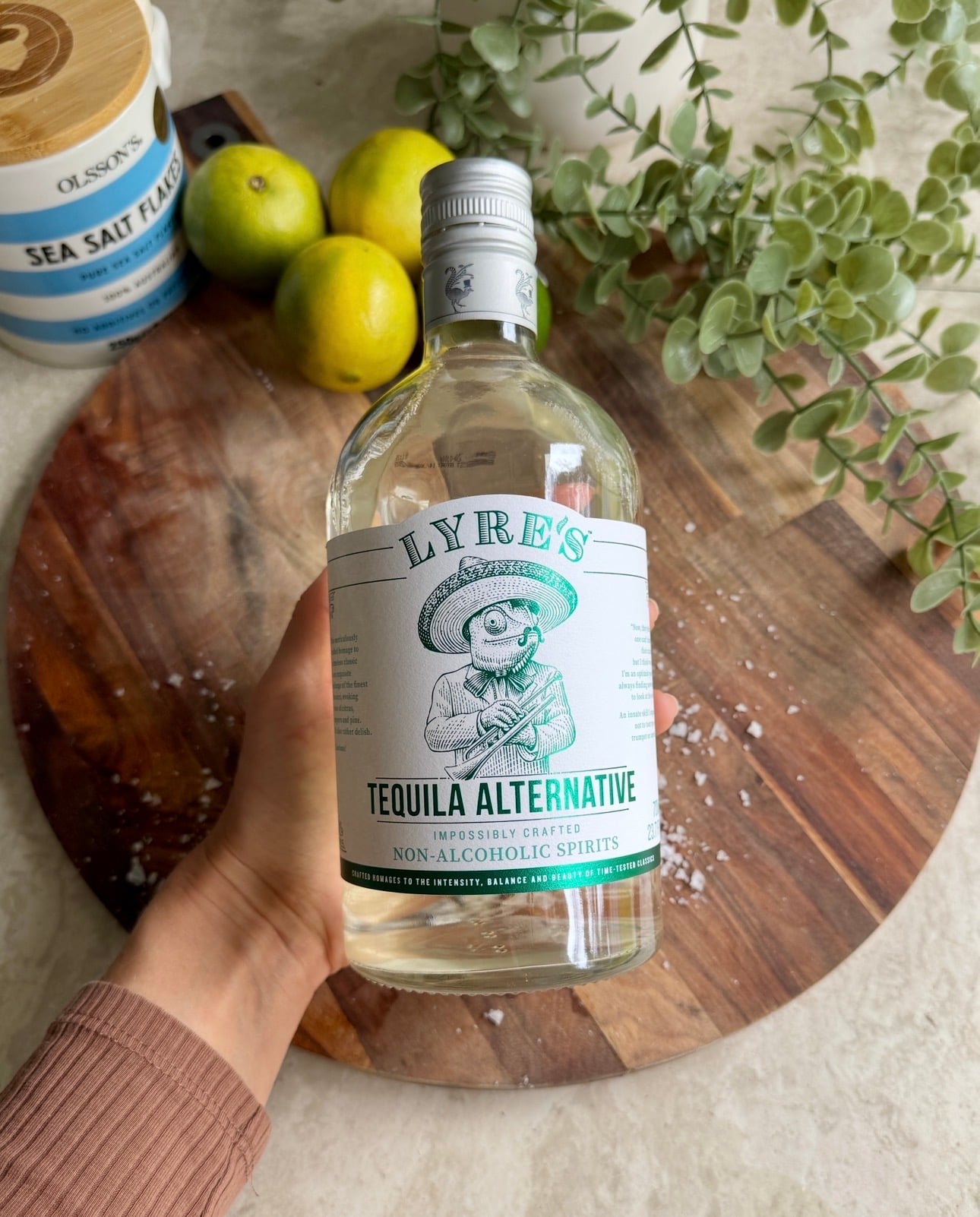 A hand holds a bottle of Lyre’s Tequila Alternative over a wooden board with limes, sea salt, foliage, and a jar of sea salt flakes in the background.