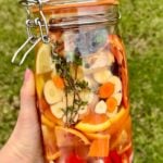 A hand holds a jar of fire cider filled with sliced vegetables, citrus, herbs, and spices, all immersed in liquid. The background is grassy and green.