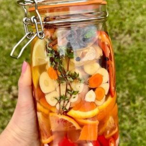 A hand holds a jar of fire cider filled with sliced vegetables, citrus, herbs, and spices, all immersed in liquid. The background is grassy and green.