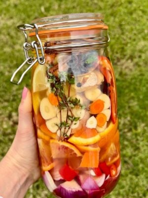 A hand holds a jar of fire cider filled with sliced vegetables, citrus, herbs, and spices, all immersed in liquid. The background is grassy and green.