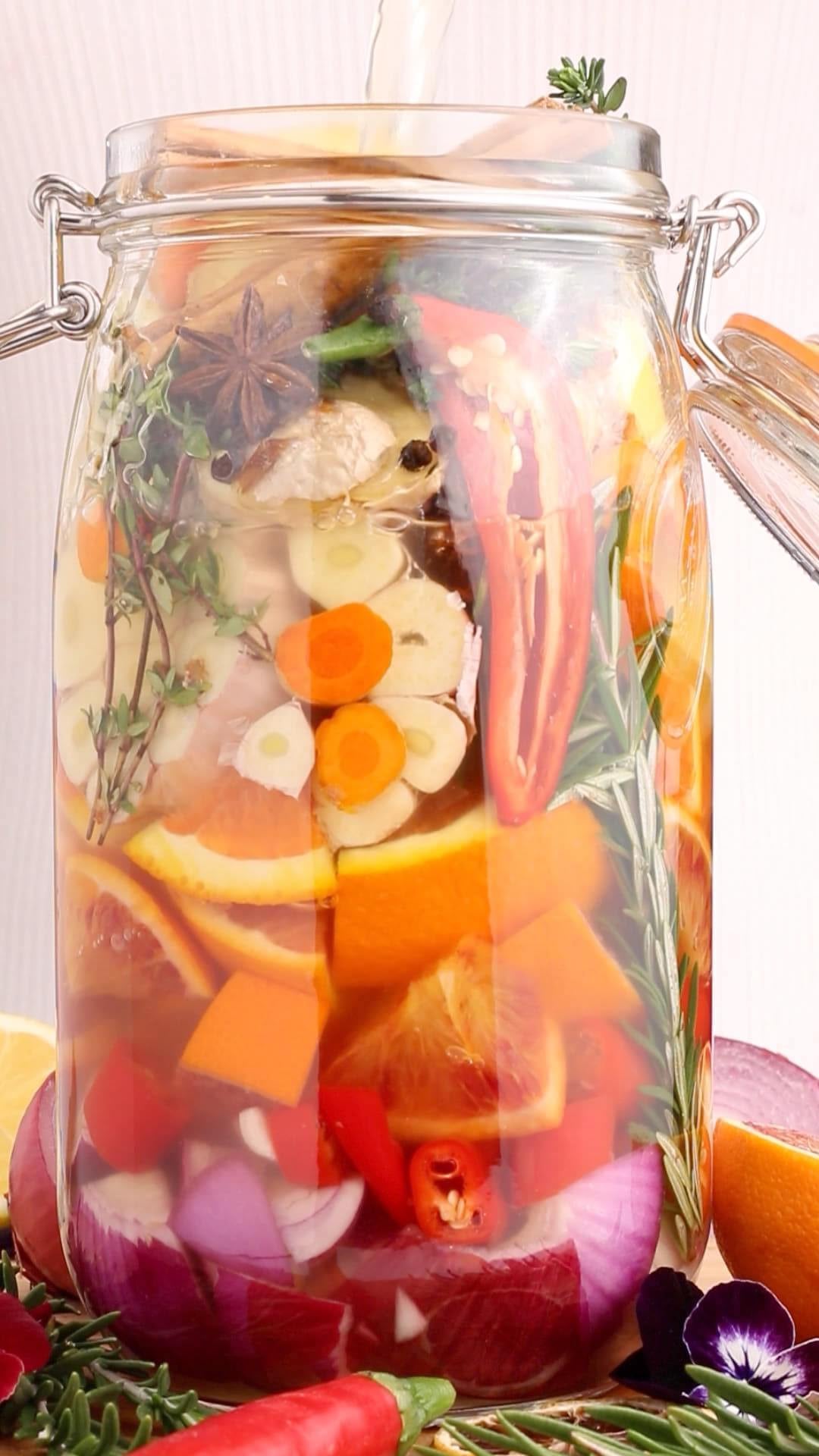 A glass jar filled with sliced oranges, garlic, chili peppers, onions, herbs, and spices, with liquid being poured in. The jar sits on a surface surrounded by fresh vegetables and flowers.