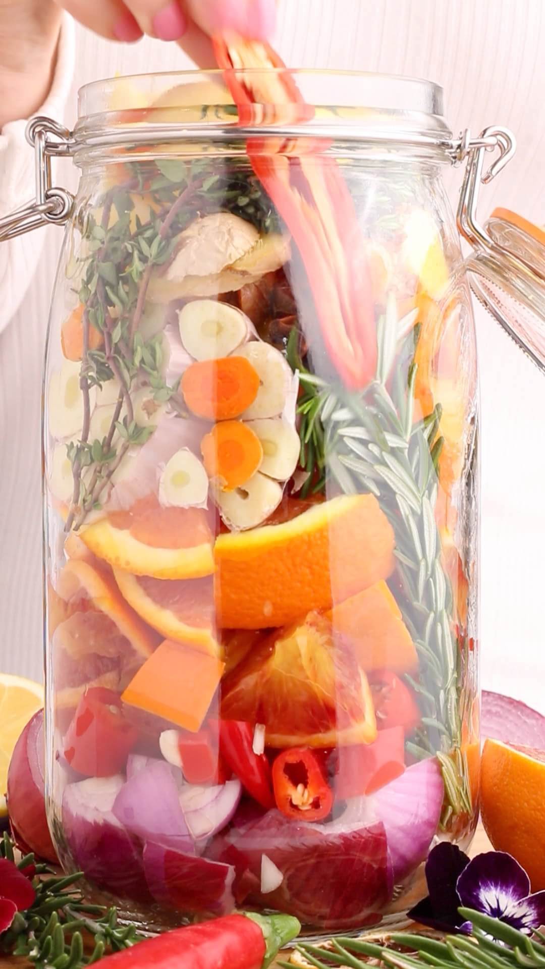 A person adds sliced carrots to a large glass jar filled with vegetables, citrus, and herbs, including oranges, onions, garlic, and rosemary.