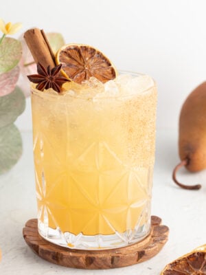 A mocktail in a patterned glass, garnished with a cinnamon stick, star anise, and a dried citrus slice, set on a wooden coaster next to pears and additional dried citrus slices.