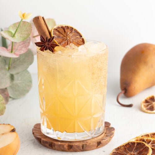 A mocktail in a patterned glass, garnished with a cinnamon stick, star anise, and a dried citrus slice, set on a wooden coaster next to pears and additional dried citrus slices.