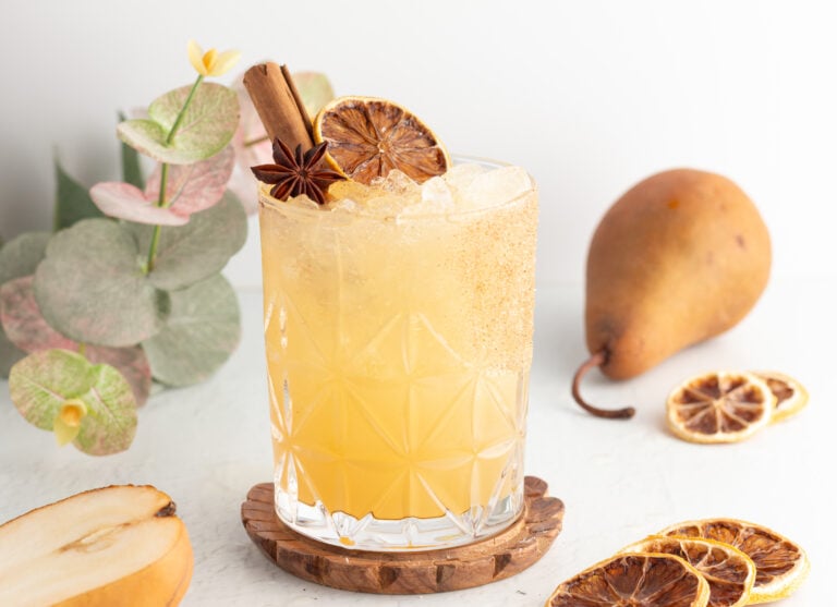 A mocktail in a patterned glass, garnished with a cinnamon stick, star anise, and a dried citrus slice, set on a wooden coaster next to pears and additional dried citrus slices.